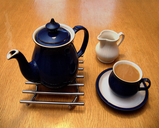 Une théière en céramique sur un dessous de plat en métal, un pot à lait et une tasse à thé pleine sur une soucoupe
